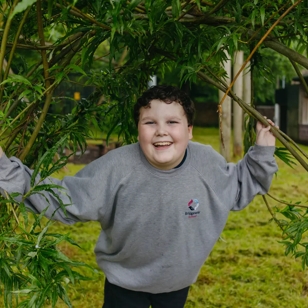 A young boy at Bridgeway School in some willows outside smiling at the camera