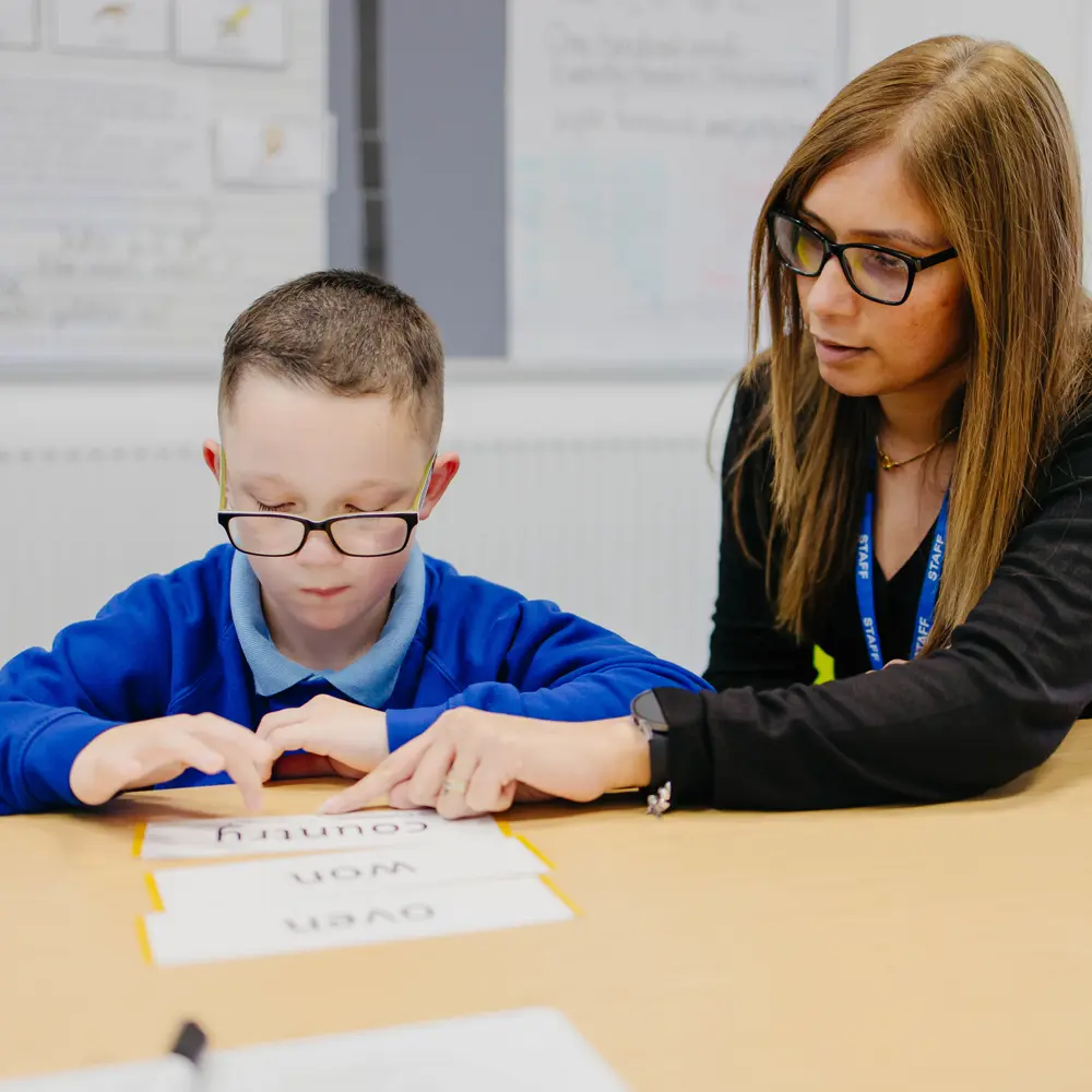 Young boy sat at a desk with teacher practicing phonics at Bescot Hall School