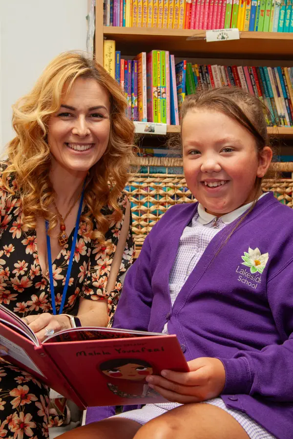 A woman and a girl smile while reading a book together in the library at Lakeside School.