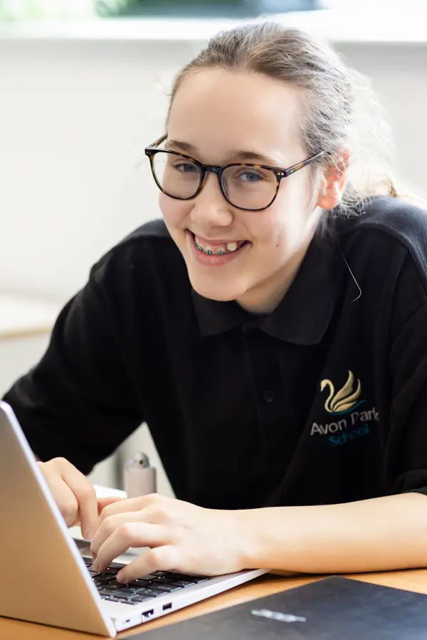 A young girl with glasses at Avon Park school smiling at the camera whilst on a laptop