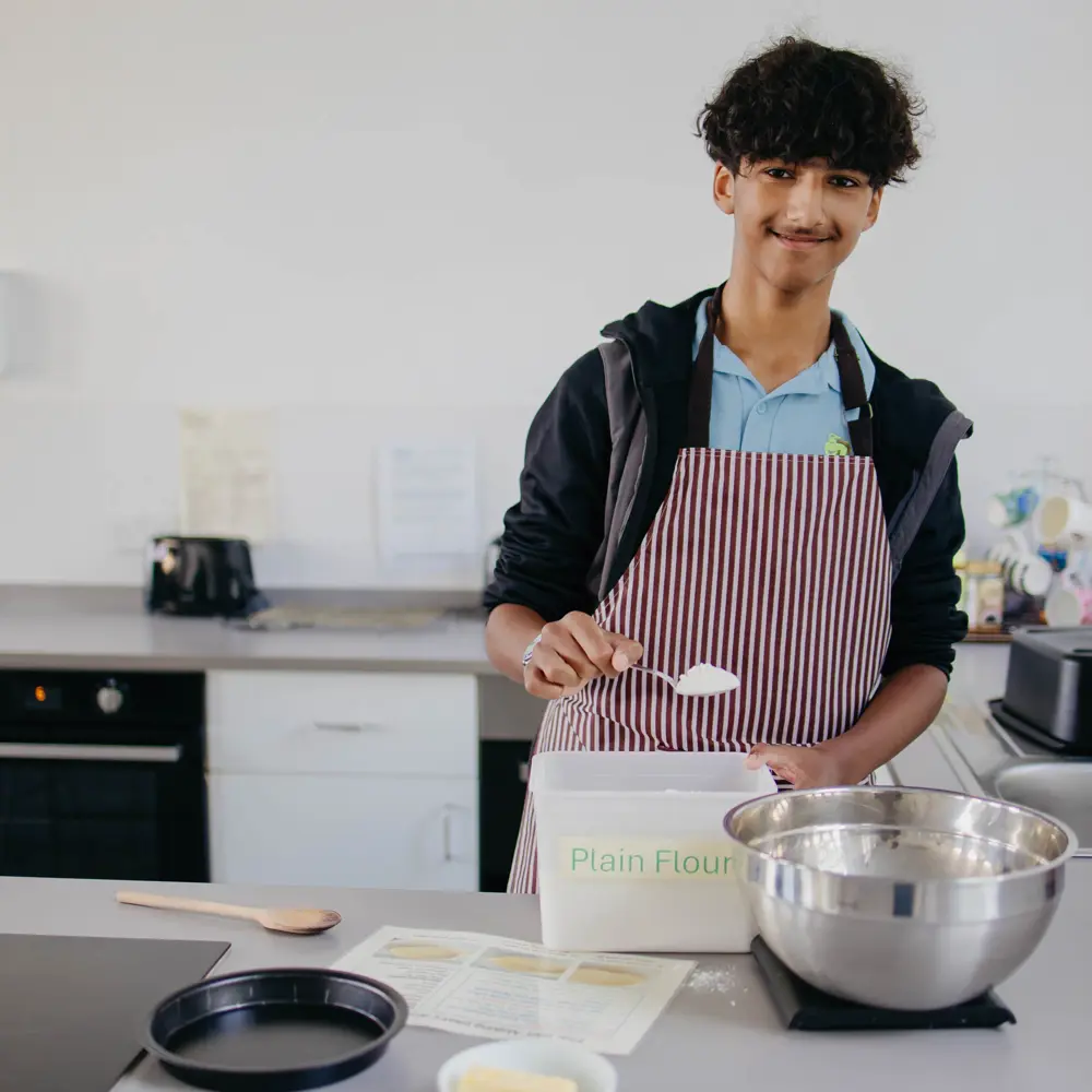 Boy in school uniform wearing a burgundy and white striped apron scooping flour in a kitchen