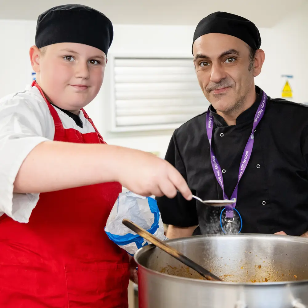A young boy and staff member cooking in a kitchen with black chef hats on at Bramley Hill