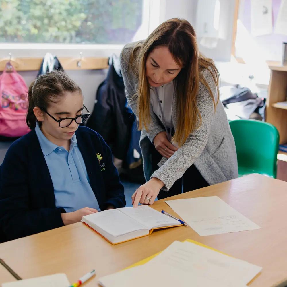 Female teacher helping a young girl with glasses in school uniform sat at a desk with her classwork