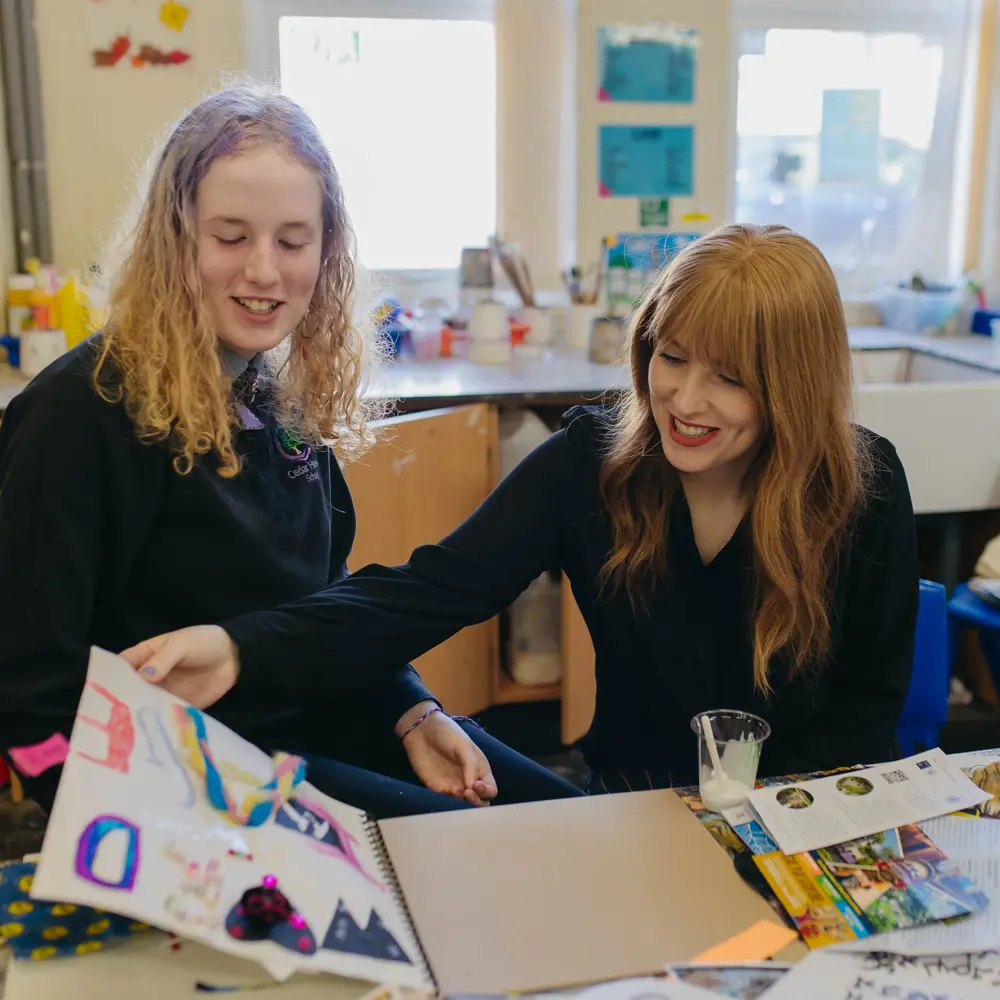 Teacher and pupil from Cedar House School sat at a desk in an art room looking through an art book